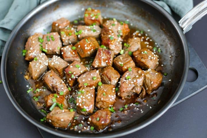 steak bites in a pan with garnishes of chives and sesame seeds.