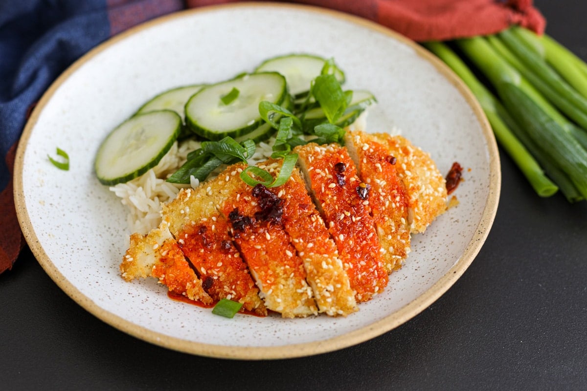 sesame chicken cutlets on a plate with rice, chili oil and cucumbers. 